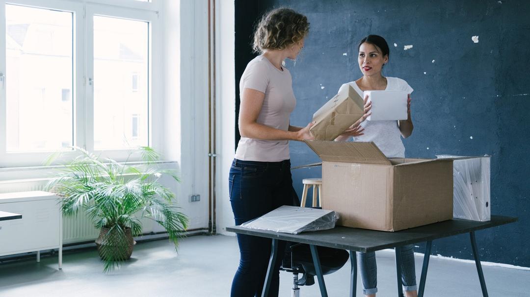 Two business women standing in an office, unpacking a box full of binders about business tax deductions.