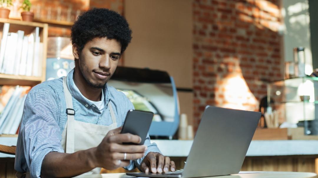 A young man wearing an apron uses a smartphone and laptop at a cafe table, with a brick wall in the background.