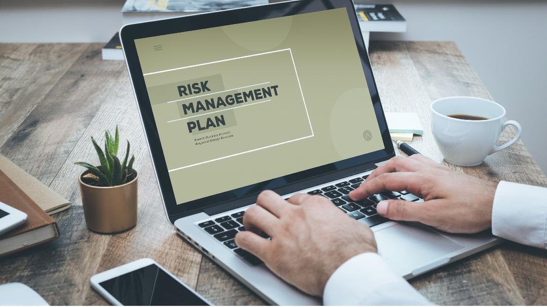 A person views a "risk management plan" on a laptop on a desk with a coffee cup, notebook, and small plant nearby.