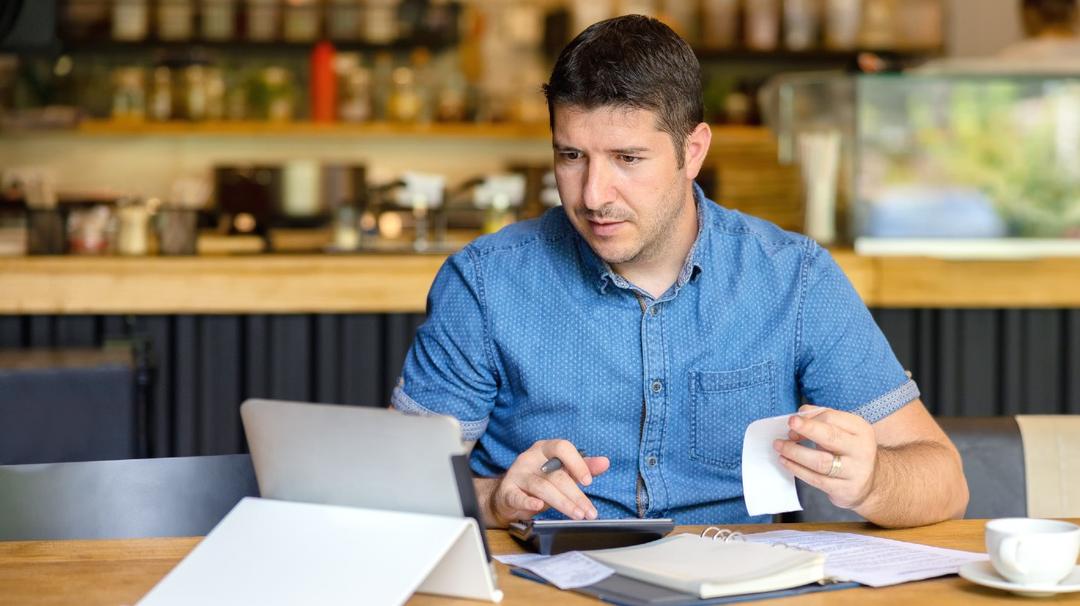 Man in blue shirt working on laptop at a coffee shop, holding a receipt and looking at the screen.