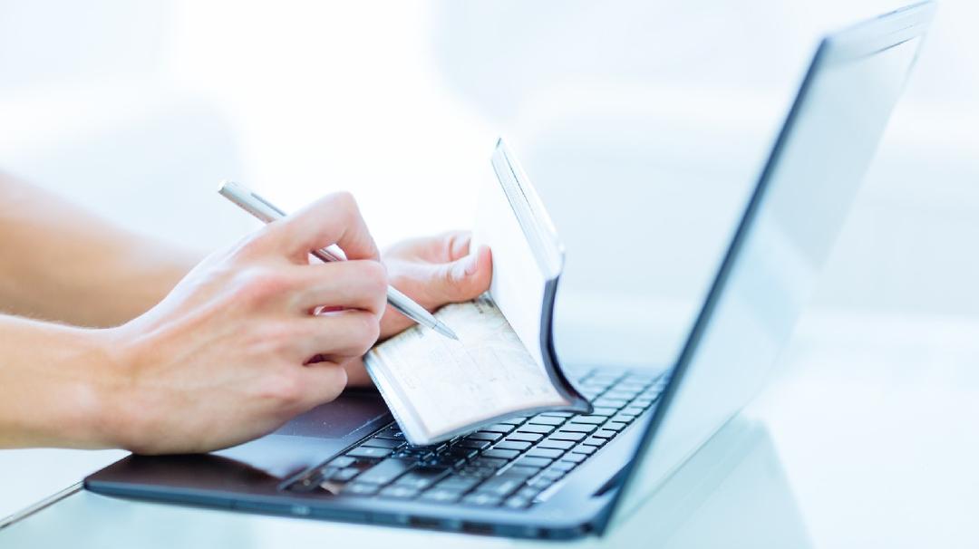 manage your bank accounts: Closeup of a caucasian male adult writing on an empty personal chequebook while using a laptop computer in a modern environment.